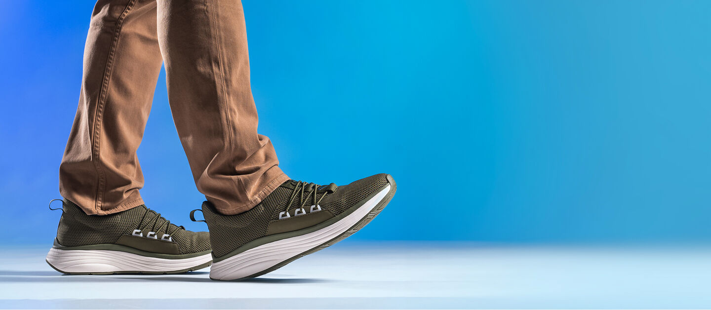 Man wearing Align Talmon in Olive with brown pants on a blue gradient background. Front foot has toe in the air.